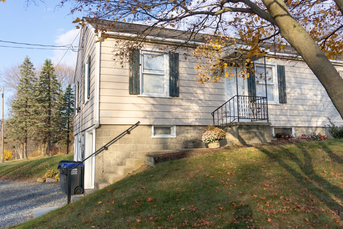 The exterior of the townhouse is depicted, showcasing a light-colored facade with green shutters. Steps lead up to the entrance, framed by seasonal foliage. A driveway with a trash bin is visible, alongside a well-maintained lawn and flower planters at the base of the building.