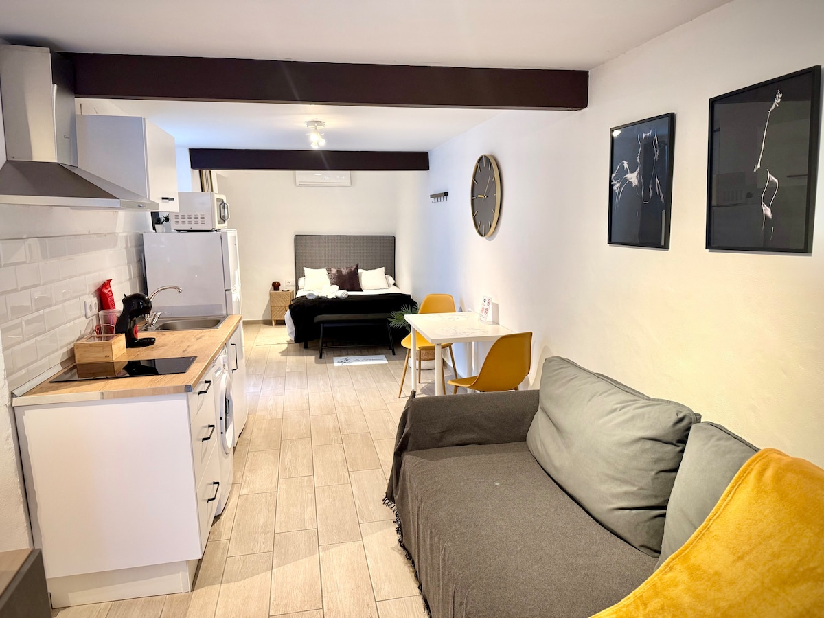 A compact living area features a cozy couch adorned with a soft throw. A wooden table with yellow chairs sits to one side. In the background, a comfortable bed is visible, accompanied by modern wall art and a wall clock, enhancing the functional layout.