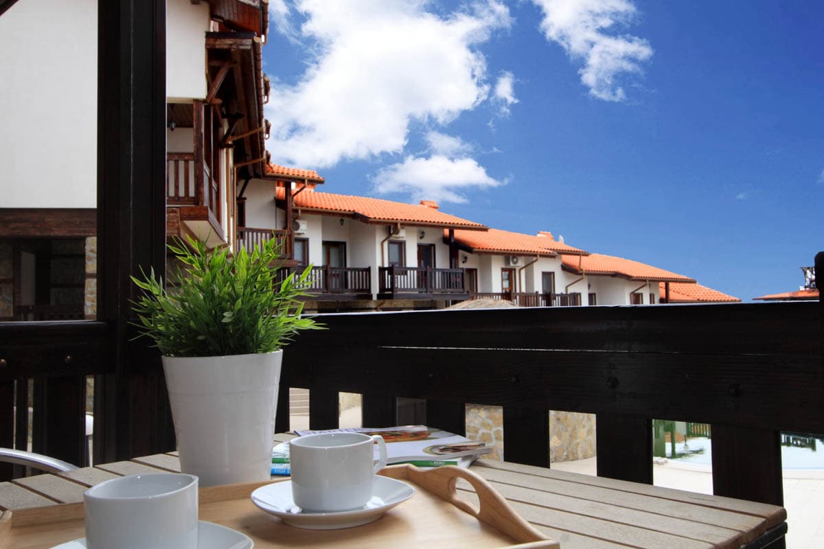 A wooden balcony is visible, featuring a light-colored tray with two cups and a small plant in a white pot. The background shows a view of traditional architecture with terracotta roofs against a clear blue sky.
