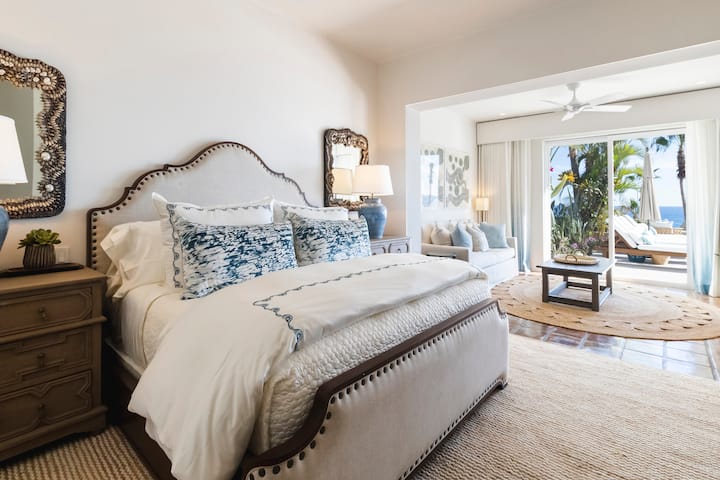 Bedroom 1 with king bed, white bedding, blue accent pillows, and dark wood nightstands. Patio doors open to a private outdoor seating area with ocean views beyond.