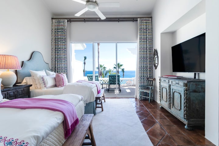 Bedroom 4 second angle showing two beds with pink accent throws, ceiling fan, flat-screen TV on rustic dresser, and patterned curtains framing sliding glass doors to an ocean-view terrace.