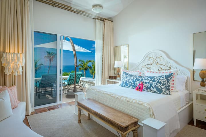 Bedroom 3 king bed with carved white headboard, blue floral pillows, and wood bench. Sliding glass doors open to ocean-view balcony with seating.