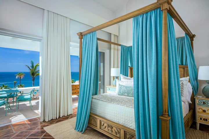 Bedroom 2 with carved wood four-poster canopy bed draped in teal silk, white bedding, and floor-to-ceiling curtains. Sliding doors open to a private terrace with direct ocean views.