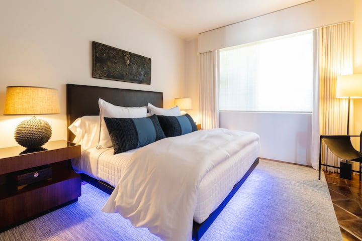 Bedroom 5 with king platform bed, dark wood headboard, navy accent pillows, white bedding, and LED under-bed lighting. Large window with floor-length drapes and warm table lamps.