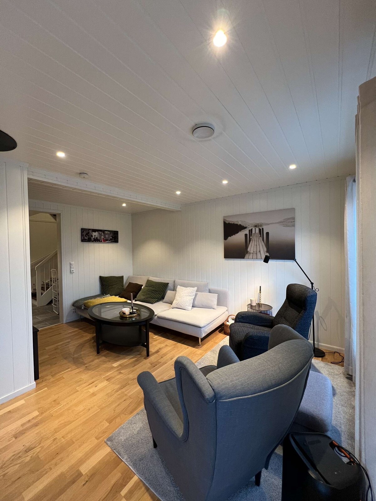 A well-lit living area features a light color palette with wooden flooring. A sectional sofa and two armchairs provide ample seating. A low round table is placed at the center. Artwork is displayed on the wall, enhancing the space's aesthetic appeal.