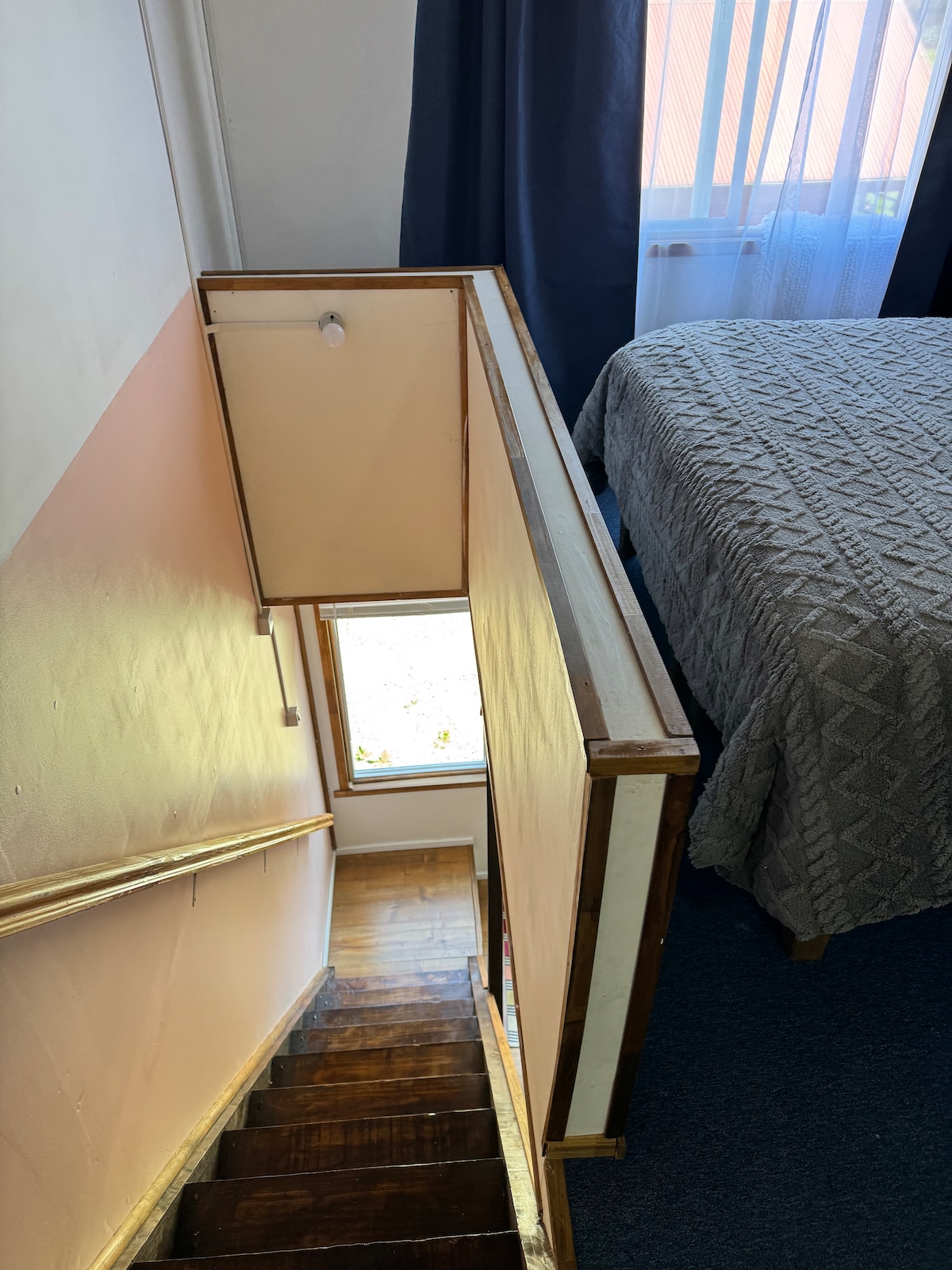 A view from the upper floor shows a staircase leading down, with warm wooden steps. The adjacent window allows natural light to fill the space, illuminating a neatly made bed with a textured blanket, partially covered by soft curtains.