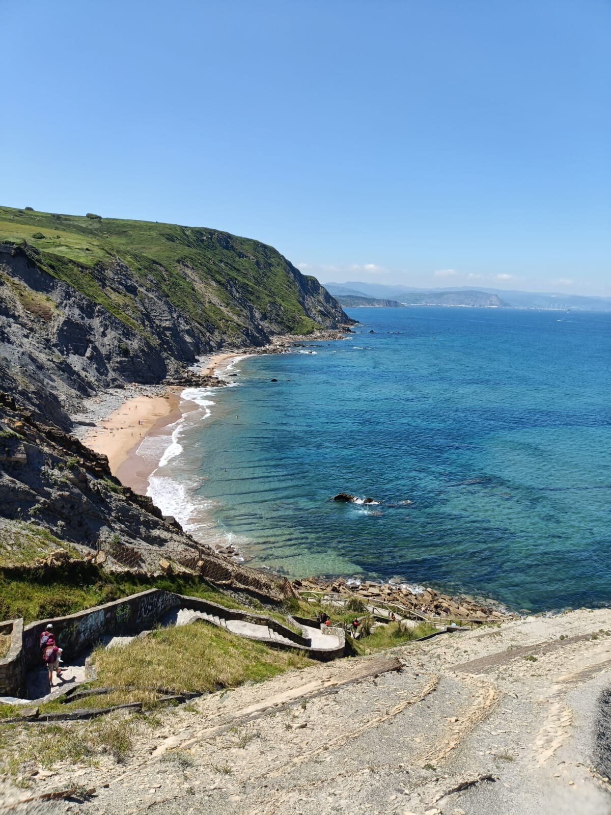 A coastal scene is displayed, featuring a sandy beach bordered by steep cliffs. Clear blue water gently laps at the shore, with gentle waves visible. The sky is bright, revealing a few distant hills on the horizon. A winding path leads down to the beach from higher ground.