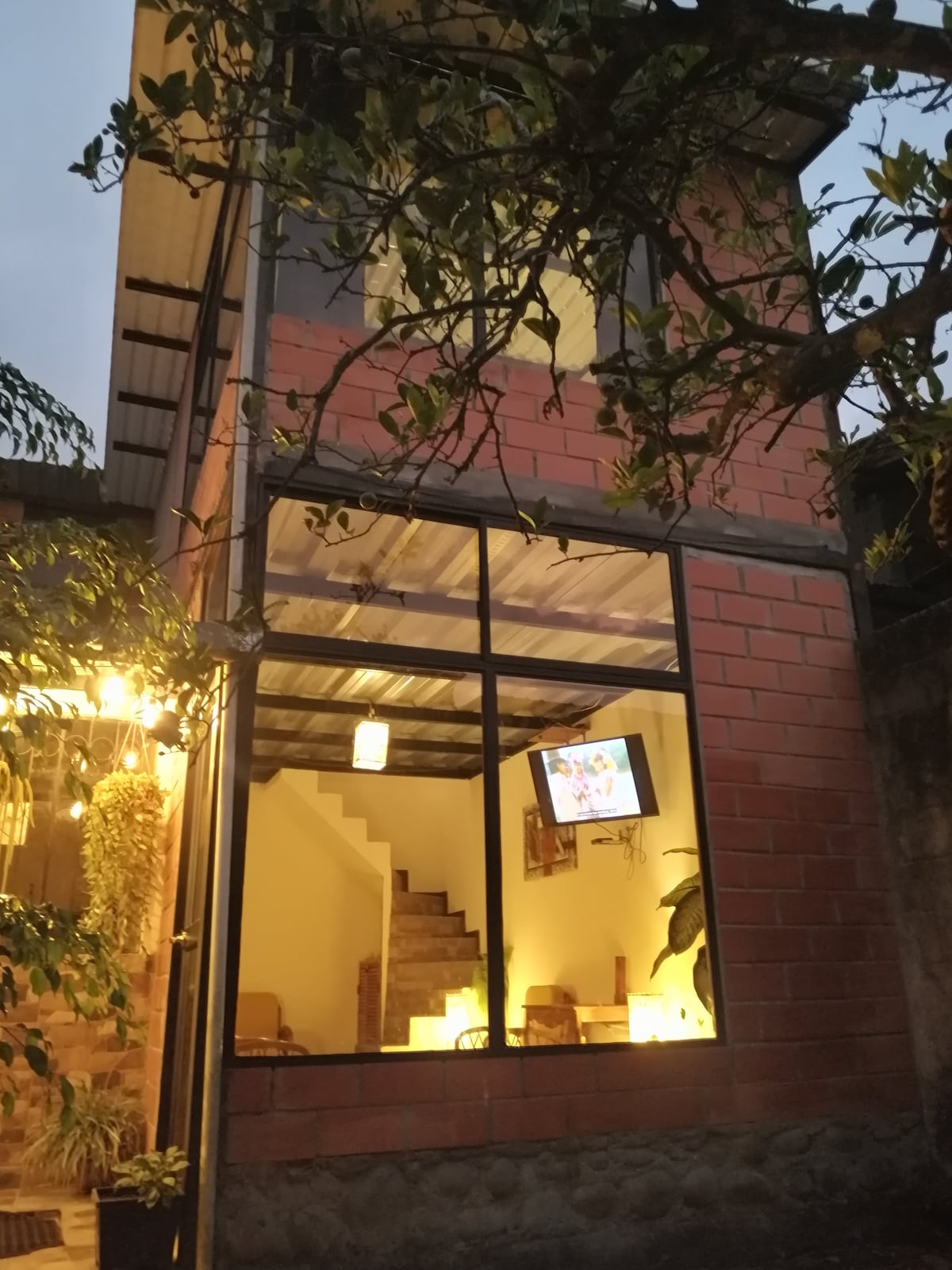 The exterior of the house is visible, showcasing a two-story structure with large glass windows. Warm lighting can be seen illuminating the interior space, where a staircase leads to the upper level. Surrounding foliage adds a natural element to the scene.