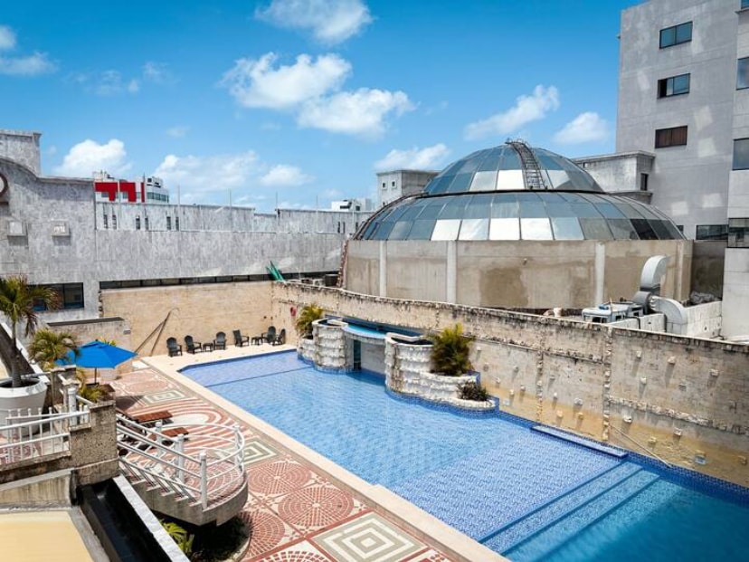 An expansive outdoor pool features a mosaic pattern and serene water features. Surrounding lounge chairs are arranged for relaxation under a clear blue sky. A distinctive dome structure is visible in the background, indicating a unique architectural design.