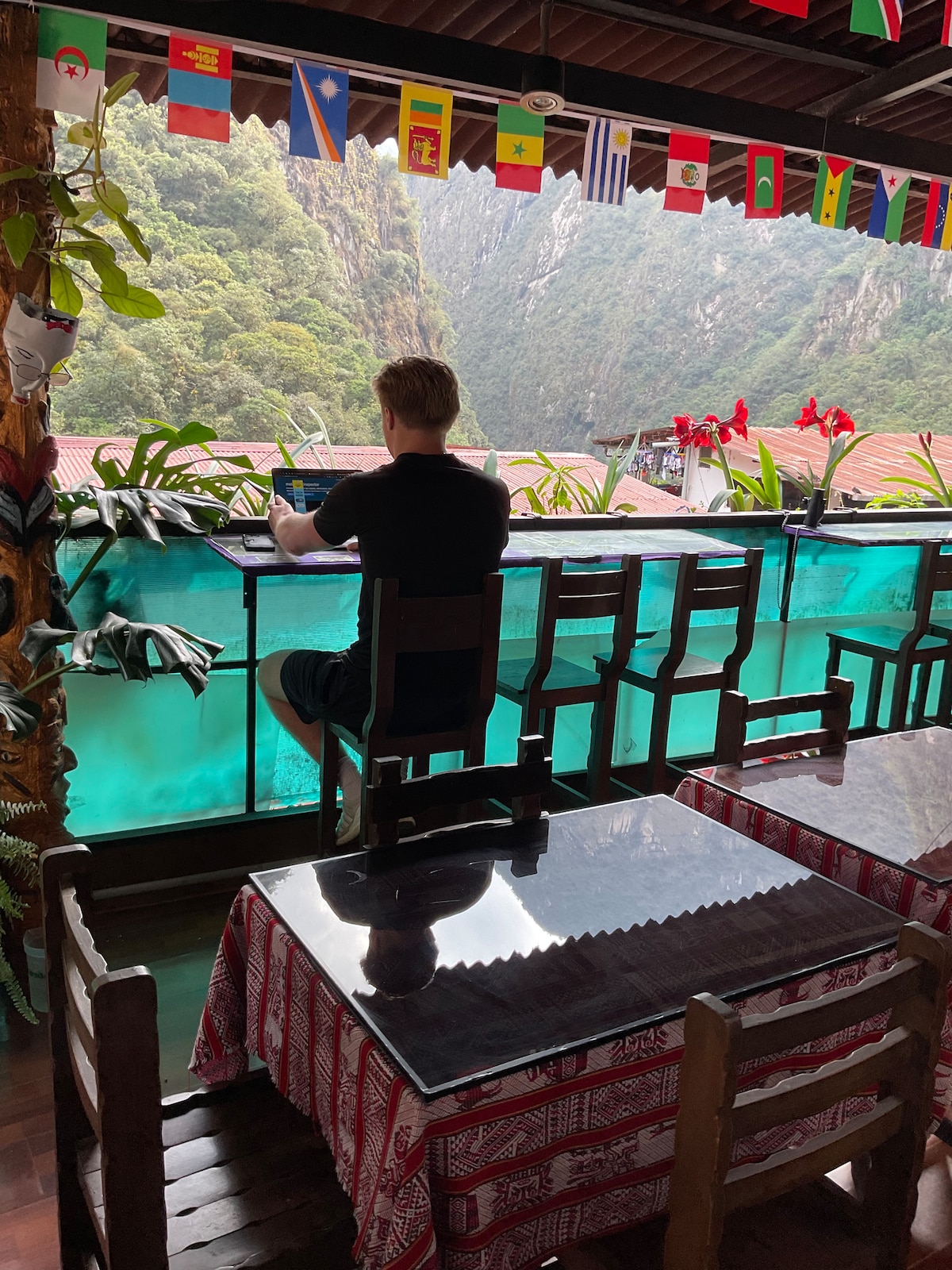 A dining area is visible, featuring wooden tables with patterned tablecloths. A patron sits on a high chair at the edge, overlooking lush green mountains through a transparent barrier. Colorful flags are displayed overhead, adding cultural flair to the serene setting.