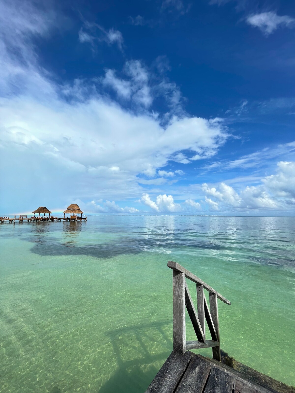 A clear view of calm turquoise waters extends towards the horizon, interspersed with fluffy clouds under a blue sky. Wooden structures with thatched roofs are visible in the distance, adding a serene element to the tranquil seascape.
