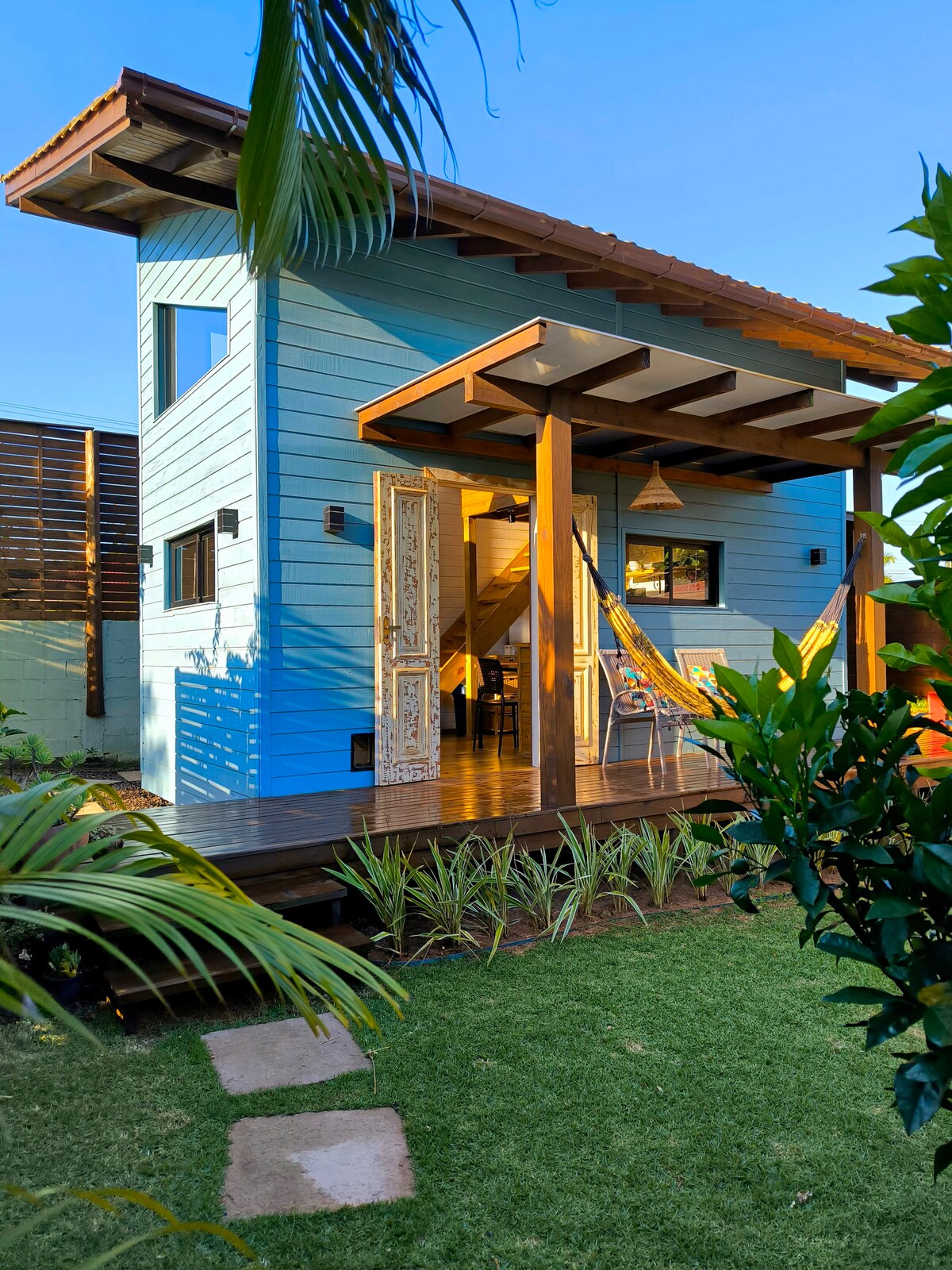 The exterior of the Tiny House features a light blue wooden facade and a welcoming porch with a hammock. The landscaped garden includes vibrant greenery and stone pathways leading to the entrance, adding to the tranquil ambiance.