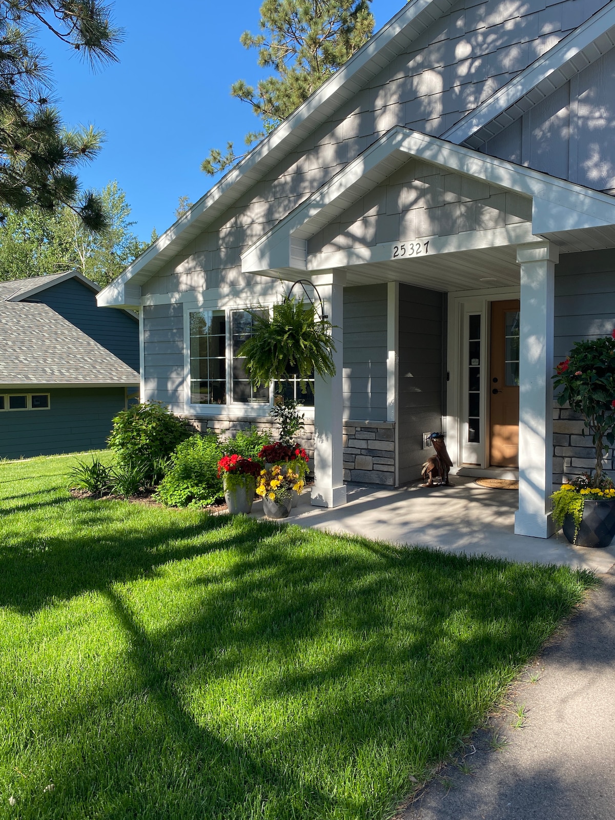 The entrance to the home features a covered porch with a welcoming ambiance. Potted plants with colorful blooms and well-maintained grass enhance the inviting setting. Soft shadows from nearby trees gently dance across the pathway, leading to the front door.