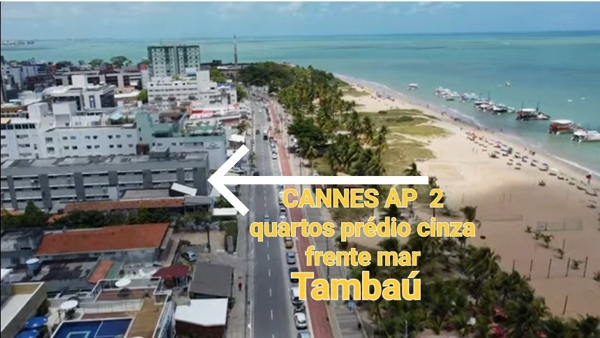 An aerial view showcases the Cannes building, marked in gray, positioned directly along the shore. A vibrant beach stretches toward the horizon, with the coastline lined by a lively road. Surrounding buildings and the serene sea are visible, contributing to the lively atmosphere of Tambaú.