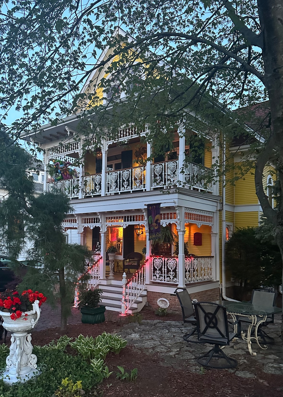 The historic Mardi Gras House displays intricate architectural details with wooden railings and decorative trim. Soft lighting from gas fixtures illuminates the exterior. The front yard features blooming flowers and outdoor seating, providing a welcoming atmosphere that draws visitors in.