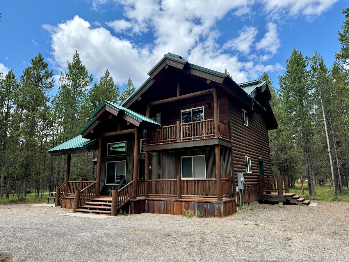 A spacious cabin featuring a rustic brown exterior and a metal roof stands amidst a scenic woodland setting. The front porch, lined with wooden railings, includes multiple staircases leading up to the entrance. Surrounding trees add natural beauty to the environment.