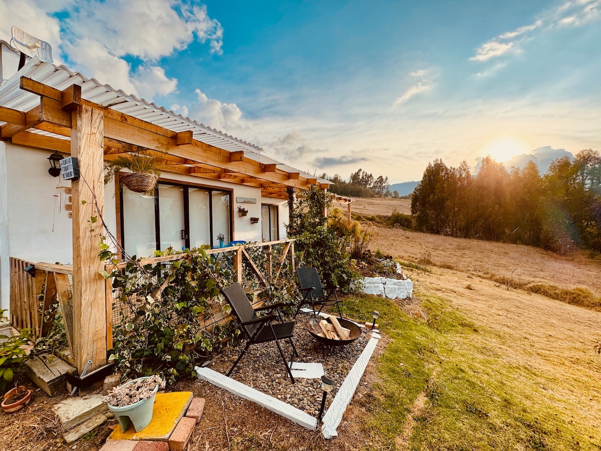 An outdoor seating area is presented with two chairs positioned near a fire pit, surrounded by greenery. The structure features wooden beams and large windows, offering views of the rolling landscape and a sunset sky filled with soft clouds.
