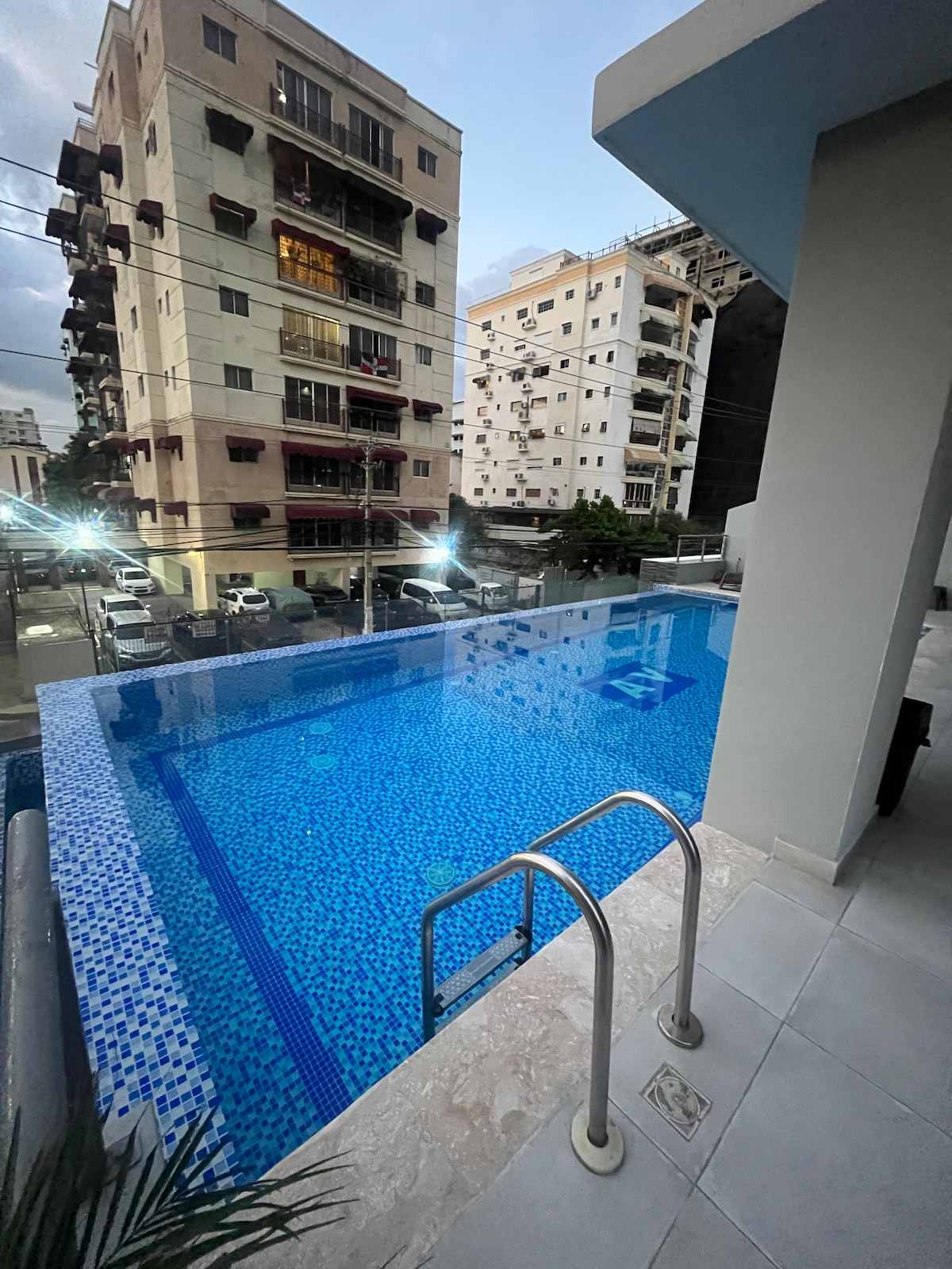 An infinity pool is visible with shimmering blue tiles reflecting the evening light. The pool's edge appears to blend into the surroundings. The area is bordered by a modern building structure and illuminated by soft lighting, enhancing a serene atmosphere for relaxation.