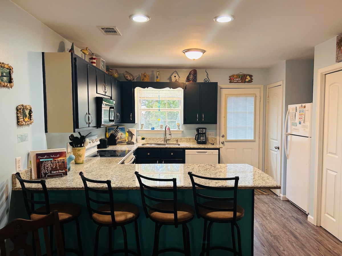 A spacious kitchen features dark cabinetry with stainless steel appliances and a large granite countertop. Barstools are arranged around the island, and a window above the sink provides natural light. Decorative items line the shelves, enhancing the inviting atmosphere.