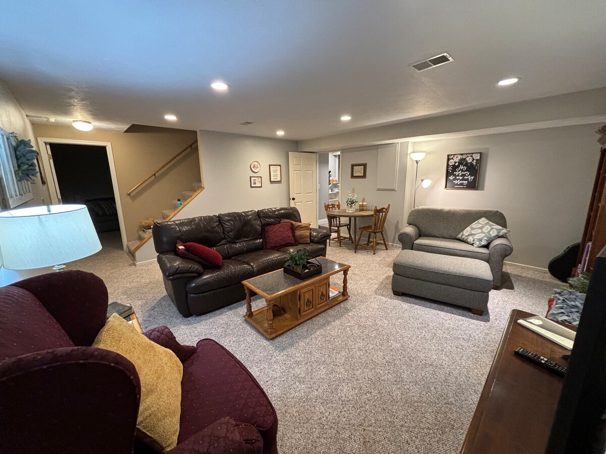 A cozy living space is seen with two distinct seating areas. A rich brown leather sofa is accompanied by a patterned armchair, while a small wooden table with chairs is positioned nearby. Soft lighting is provided by floor lamps, illuminating the carpeted floor and walls.