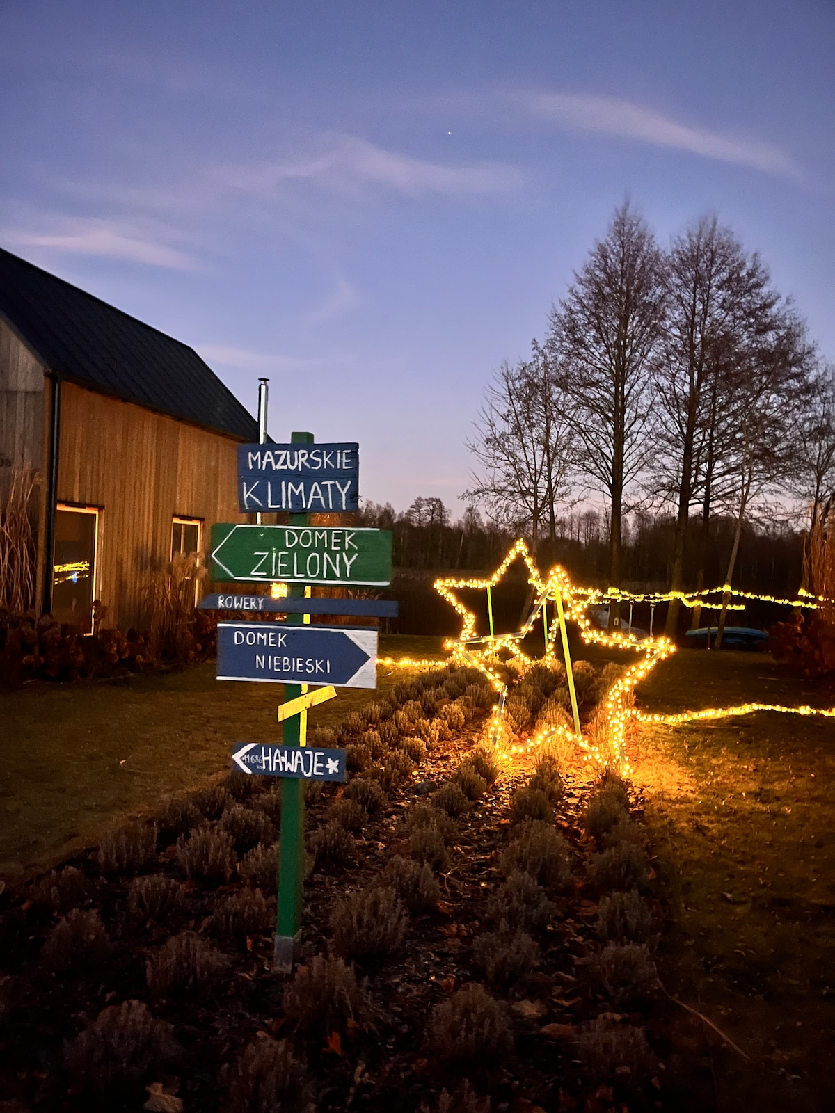 A collection of directional signs is displayed in a garden, illuminated by string lights. A star-shaped decoration is visible, enhancing the inviting atmosphere. The background features a wooden house and softly lit trees, set against a twilight sky.