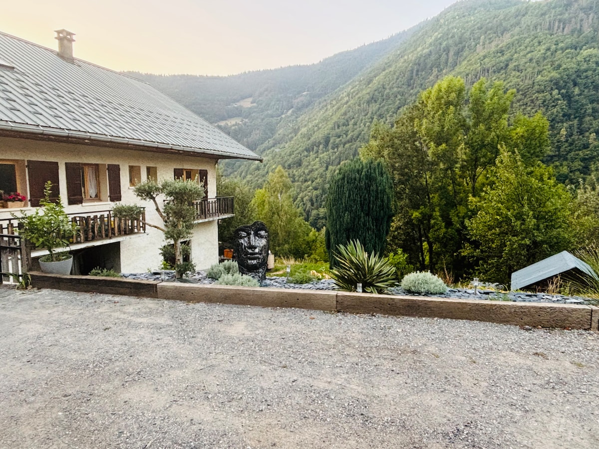 A gravel path leads towards a rustic building surrounded by lush greenery and mountainous terrain. A large stone statue can be seen in the garden area, accompanied by various plants. The serene environment is complemented by the gentle slope of the hillside in the background.