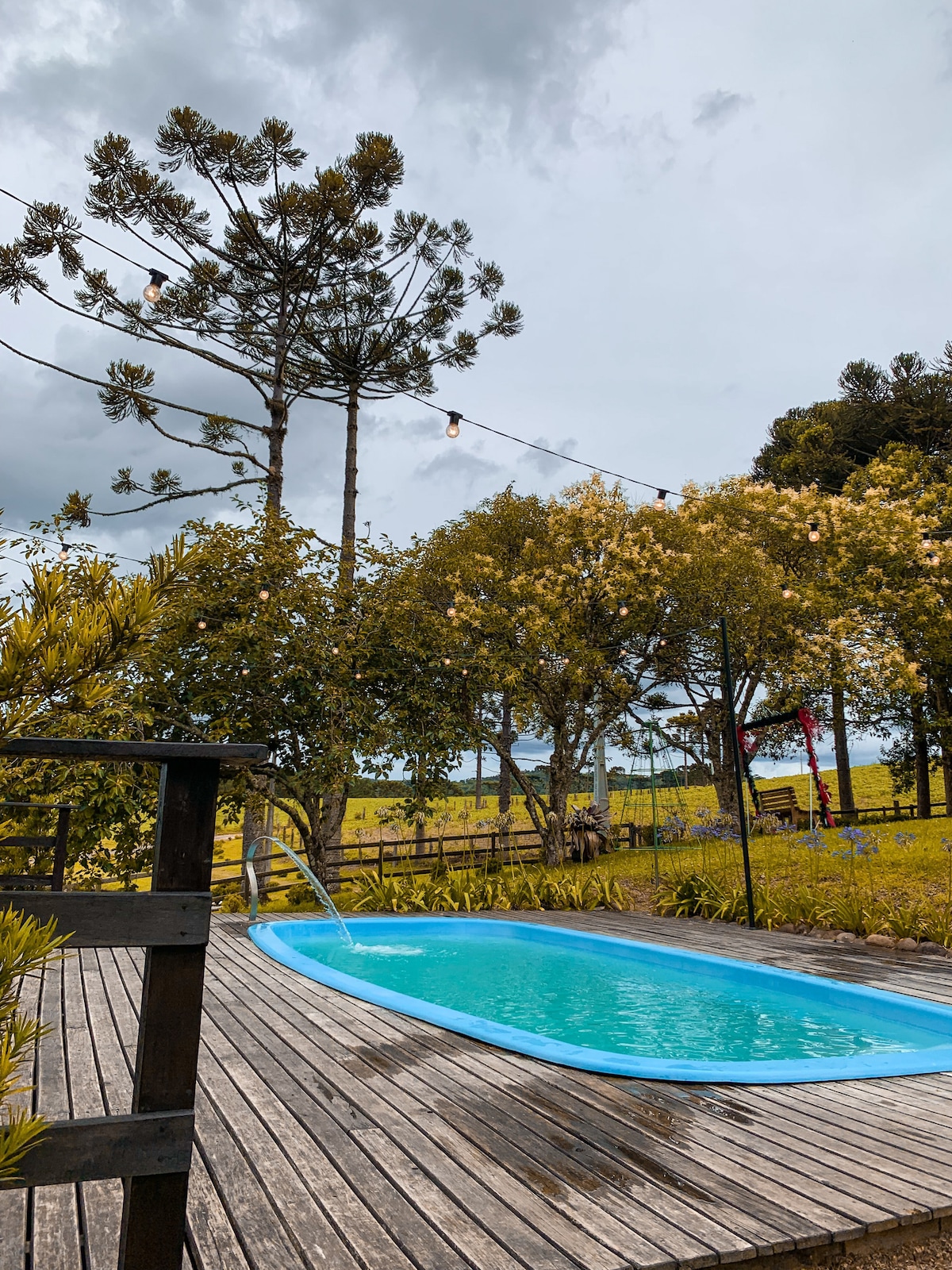 A small swimming pool is highlighted on a wooden deck, surrounded by lush greenery and blooming trees. Overhead lights are strung across the area, creating a cozy ambiance. The background features a panoramic view of open fields under a cloudy sky.