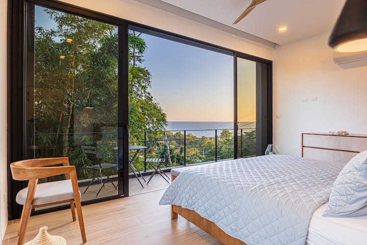 Chambre principale paisible avec un très grand lit, des portes coulissantes du sol au plafond et une terrasse privée donnant sur la jungle luxuriante et l'océan. Une retraite paisible et confortable conçue pour l'intimité et la détente.