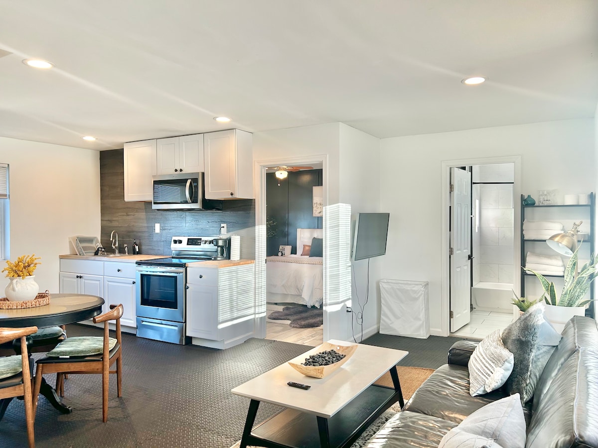 An open living space is showcased, featuring a sleek kitchen with modern appliances, a dining table with four chairs, and a comfortable seating area. Natural light brightens the area, complemented by neutral tones and simple decor. A full bathroom is visible through a doorway.