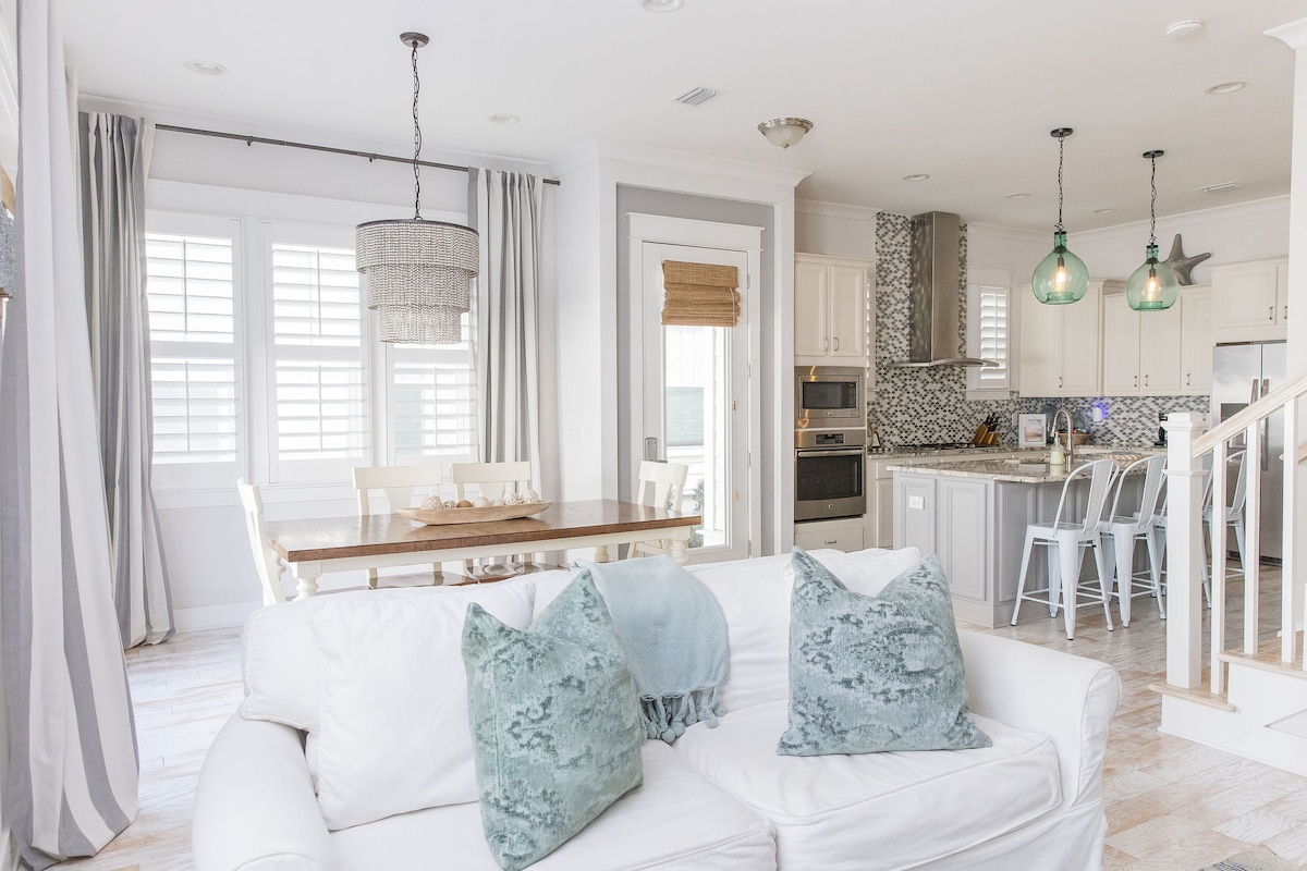 An open-concept living area is highlighted, featuring a comfortable white sofa adorned with teal accent pillows. A wooden dining table is positioned nearby, complemented by stylish pendant lighting above. The kitchen is visible in the background, showcasing modern appliances and a textured backsplash.