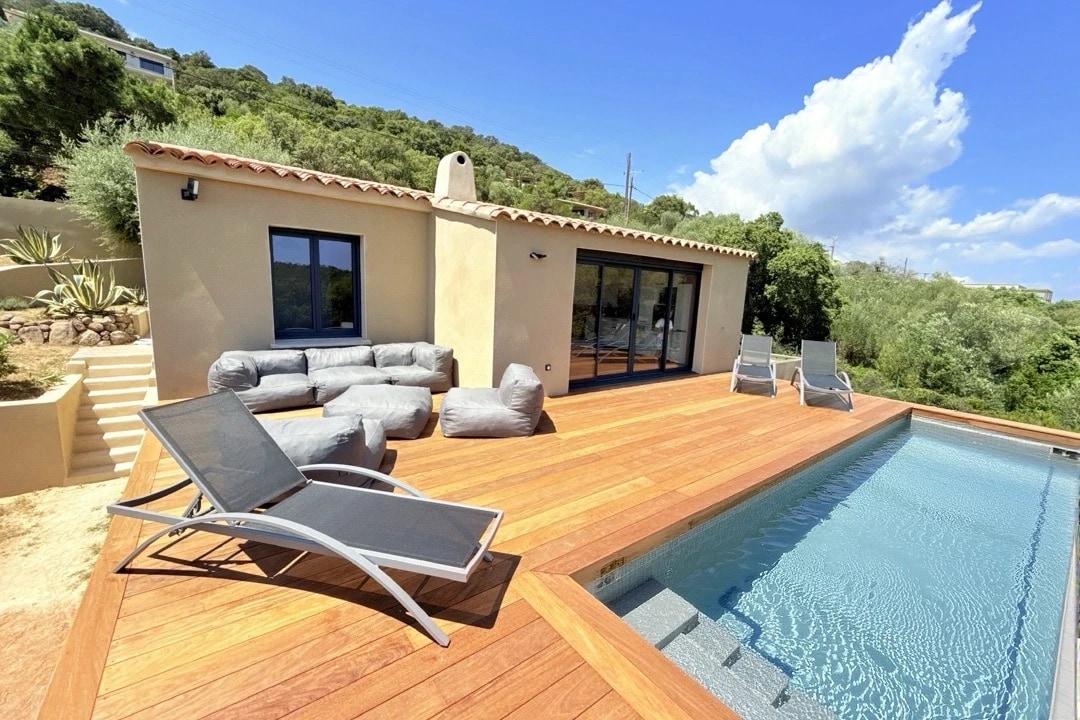A spacious wooden deck surrounds a private swimming pool, featuring two comfortable loungers positioned for relaxation. The exterior of the house is visible with large glass doors offering views of the landscaped area with greenery, and the sky displaying a few clouds.
