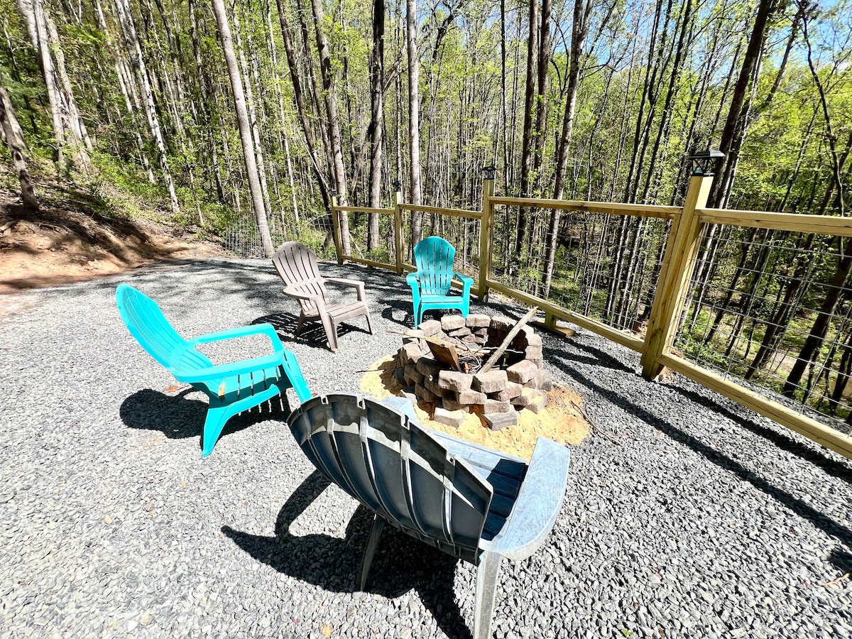 A gathering area features a firepit surrounded by a mix of seating, including two blue chairs and two gray chairs. The space is set on gravel, with a wooden fence providing a backdrop of trees, enhancing the inviting outdoor atmosphere.