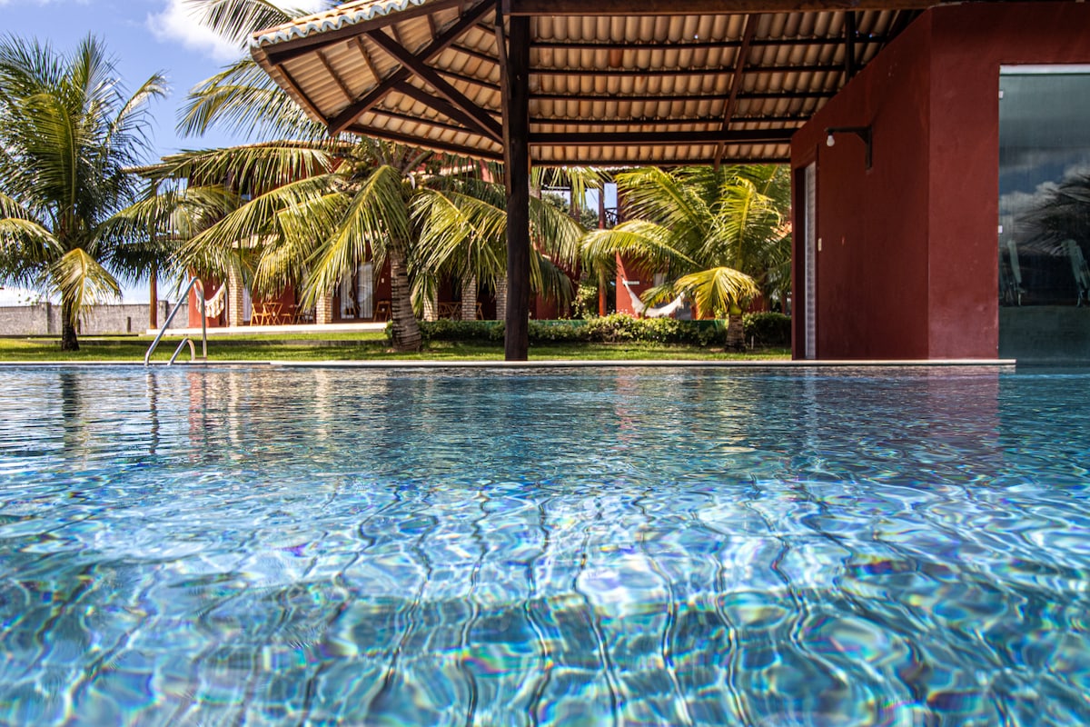 The image showcases a tranquil swimming pool with clear water, reflecting sunlight. In the background, palm trees can be seen alongside a covered lounging area, providing shade and a relaxing atmosphere. The pool's edge merges seamlessly with the surrounding landscape.