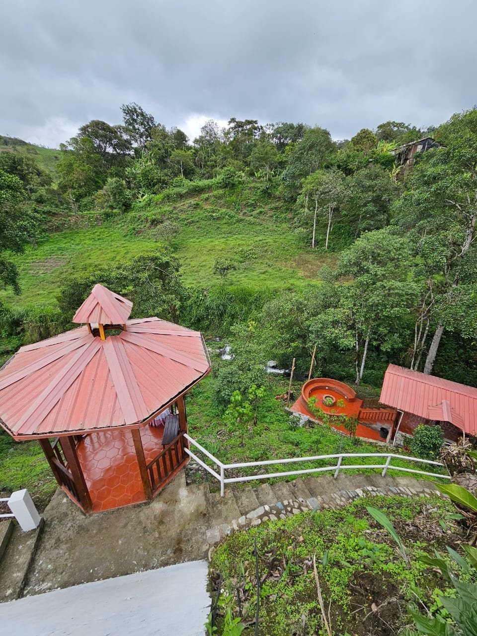 A red-roofed gazebo is visible, set among lush greenery. A slide is positioned nearby, complementing the natural landscape. The area is characterized by diverse trees and slopes, creating a peaceful environment perfect for relaxation and play.