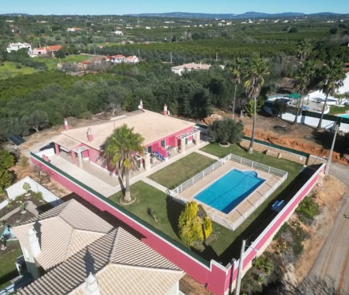 An aerial view captures the expansive property surrounded by lush greenery, featuring a rectangular saltwater pool framed by a garden. The vibrant exterior of the building contrasts with the landscape, which includes palm trees and rural scenery, illustrating a tranquil rural setting.