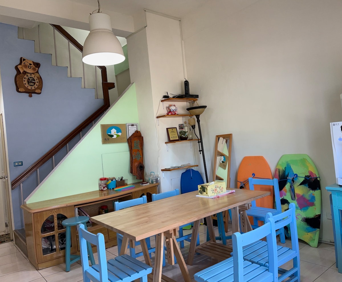 A bright dining area features a wooden table surrounded by blue chairs. A staircase leads to an upper level, with a bear-themed decoration on the wall. Colorful surfboards are positioned against the wall, adding a playful touch to the space.
