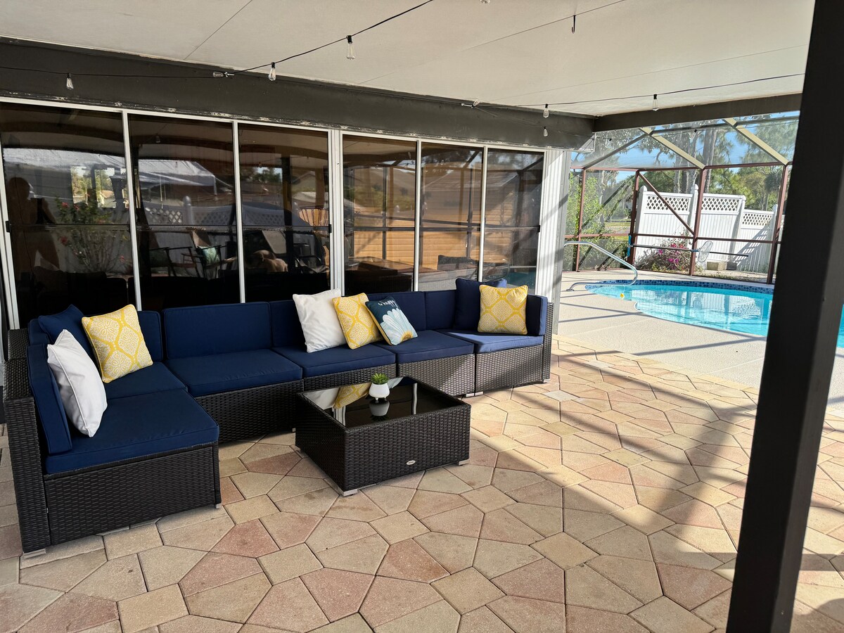An outdoor seating area features a deep blue sectional sofa adorned with white and yellow cushions. A glass-top coffee table is positioned in front, surrounded by pavers. The space is enclosed by screens, providing views of the heated saltwater pool visible in the background.