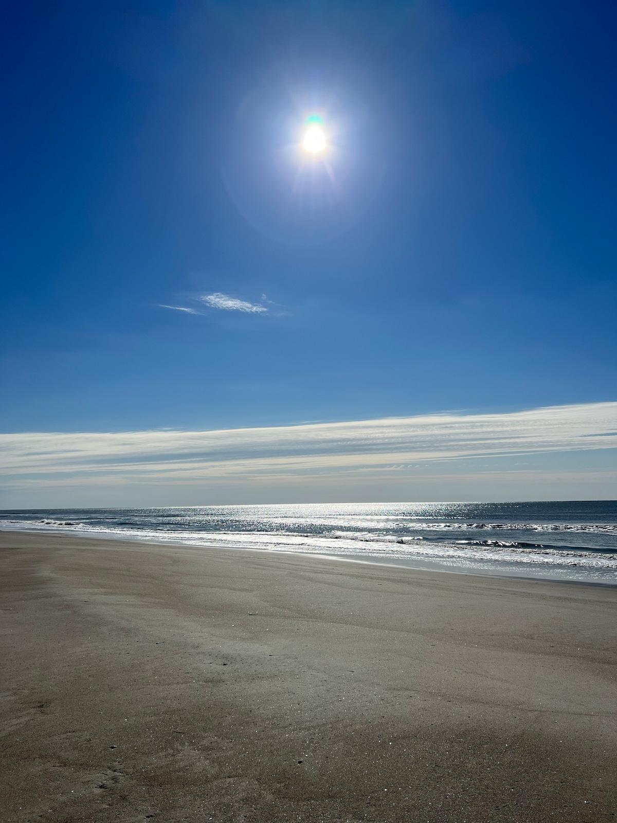 A serene beach scene is displayed, featuring fine sandy shores extending toward gentle ocean waves. The bright sky is partially adorned with soft clouds, while sunlight glistens on the water's surface, creating a calm and inviting atmosphere.