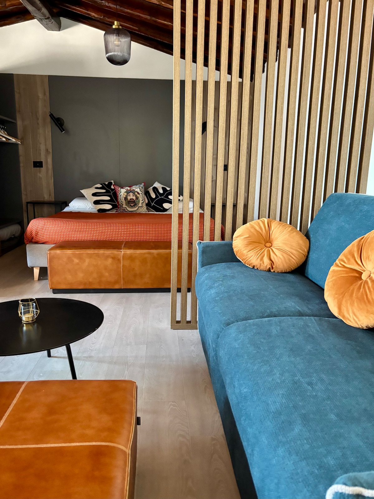 A contemporary living space features a comfortable blue sofa with orange cushions, paired with a small round table. A wooden screen partially divides the room, revealing a cozy sleeping area with a spacious bed adorned with colorful bedding.