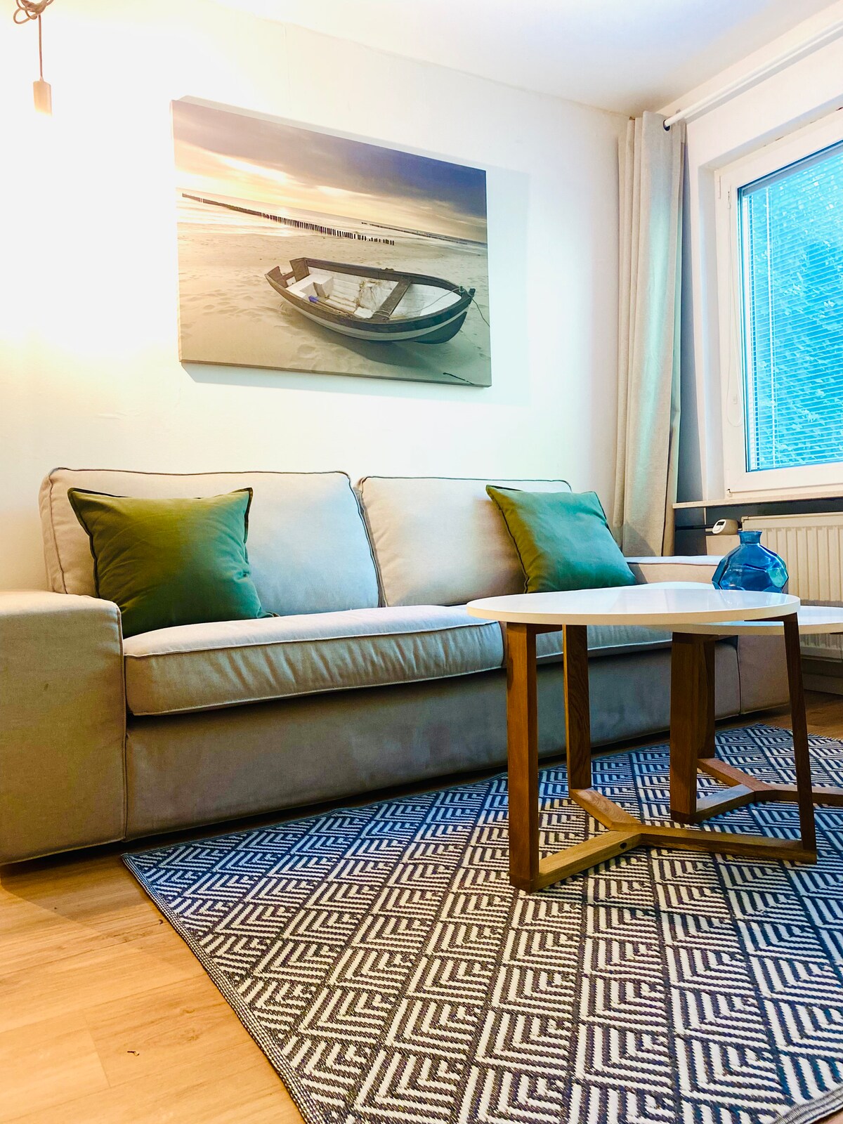 A comfortable sofa adorned with green cushions is positioned beside a wooden side table. A decorative blue bottle sits on the table. Natural light filters through a window with light curtains, and a canvas depicting a boat on the beach hangs above the sofa.