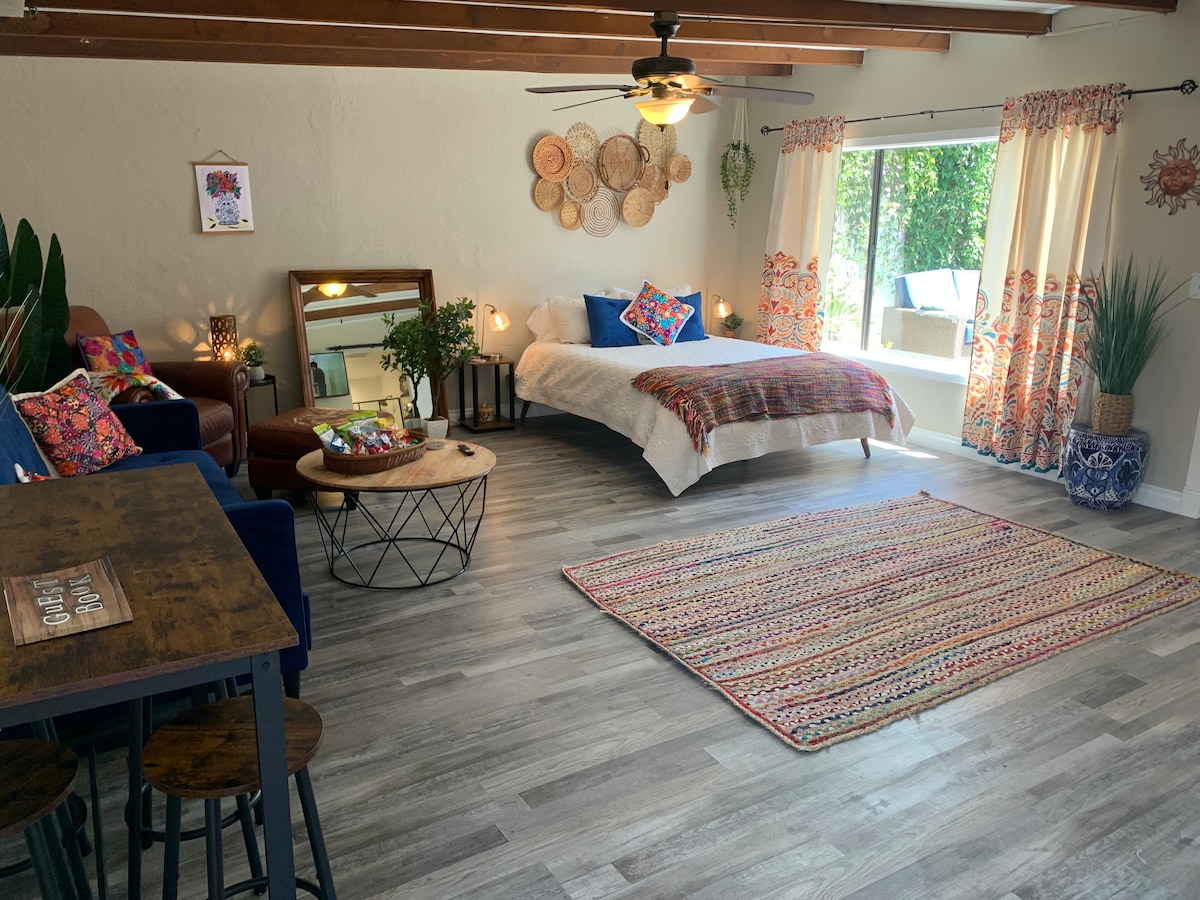 The spacious single room features a queen bed with colorful throw pillows and a textured blanket. A sitting area is offered with blue chairs and a wooden table. Natural light floods through large windows adorned with patterned curtains, illuminating a woven area rug on the floor.