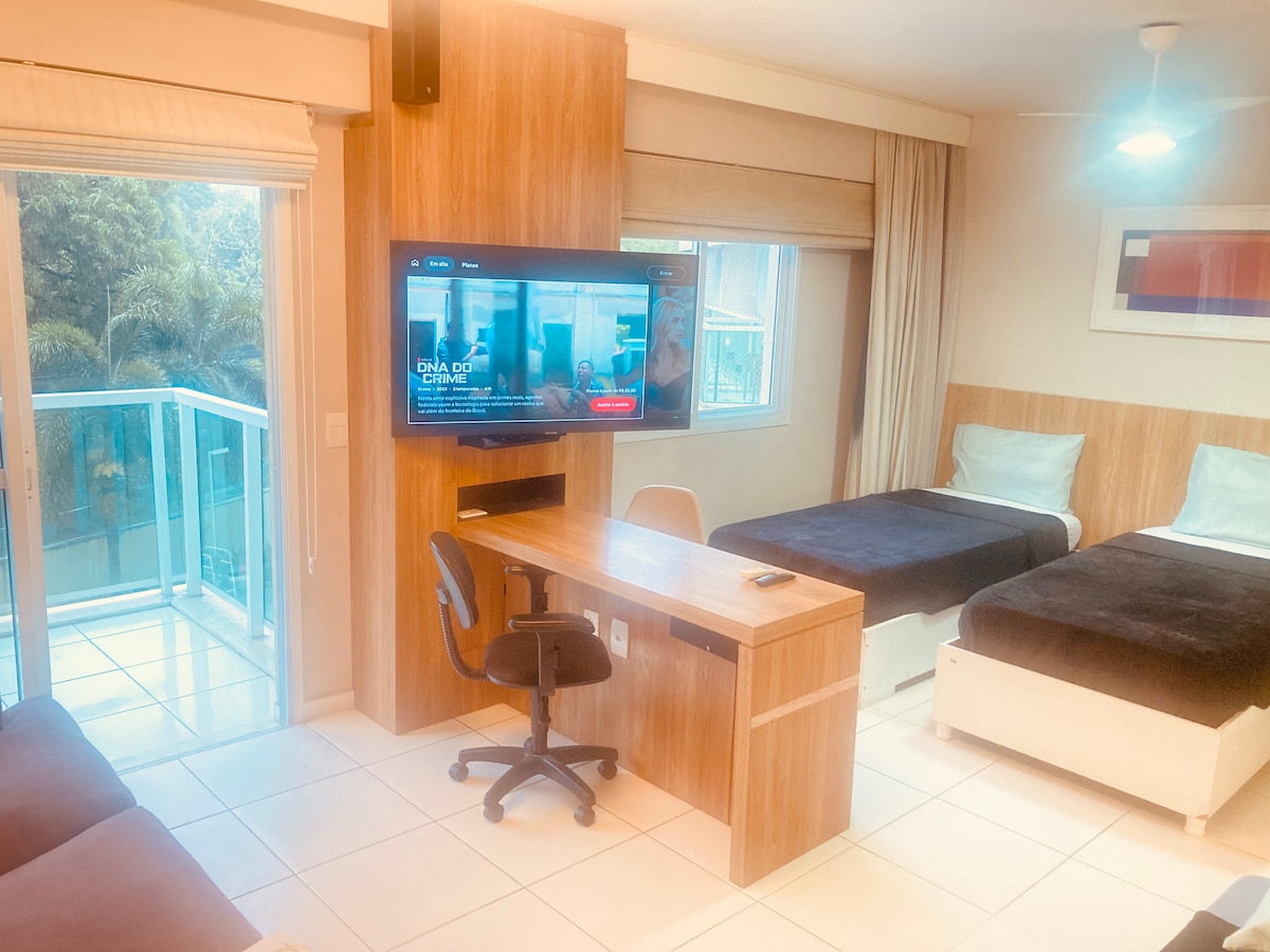 A modern room features two twin beds positioned beside a stylish work desk equipped with a comfortable chair. A wall-mounted television is positioned above the desk, and large windows allow natural light to illuminate the space. A small balcony is accessible, offering views of greenery.