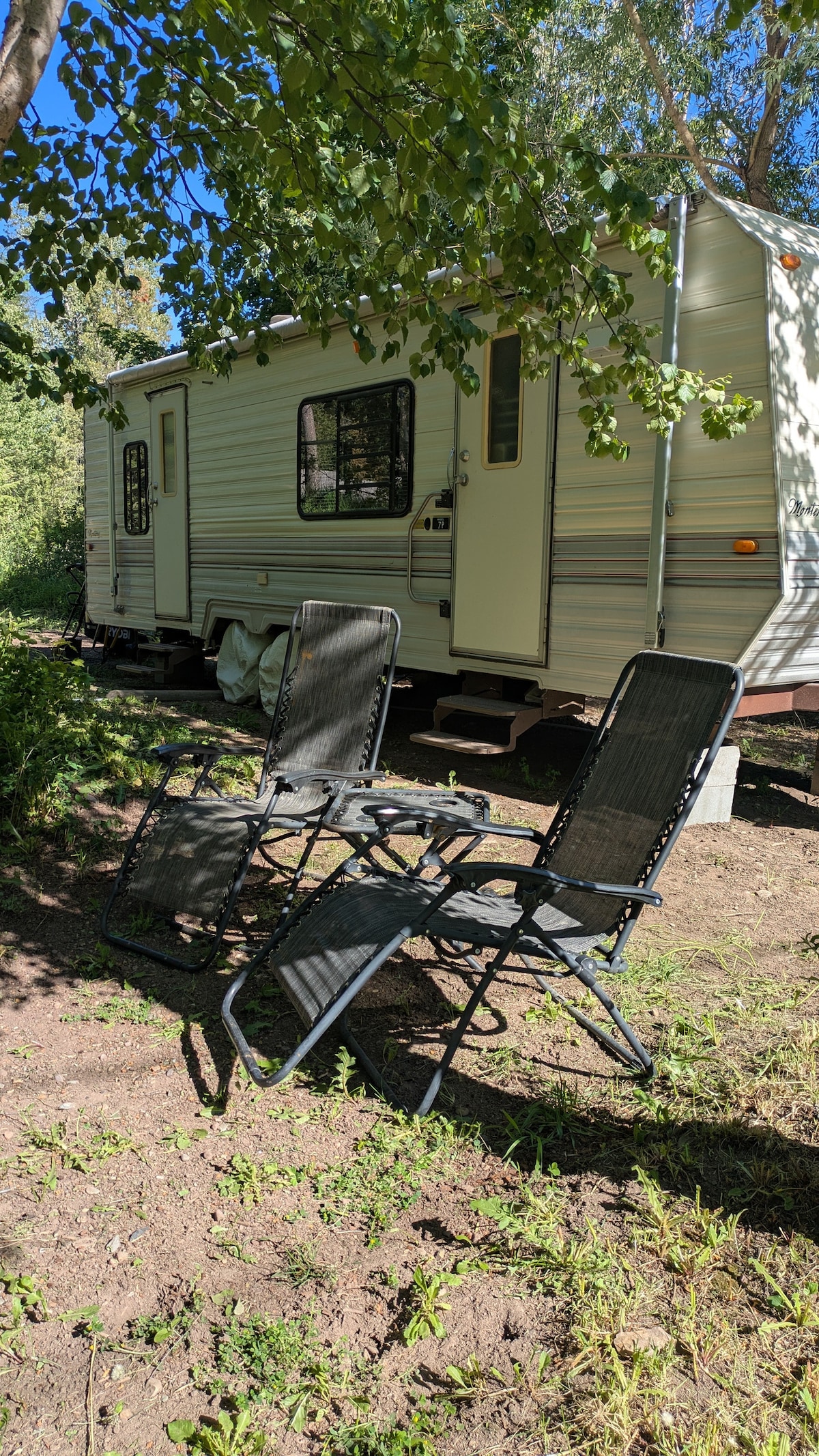 Two adjustable black chairs are positioned on a grassy area, creating a space for relaxation. The vintage camper is visible in the background, surrounded by trees, with sunlight filtering through the leaves, enhancing the outdoor setting.