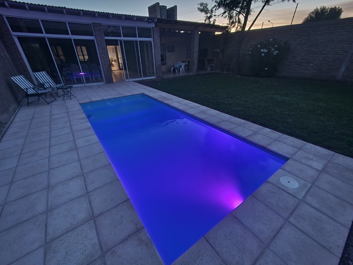 A rectangular pool with gently illuminated water casts a calming hue. Sun loungers are positioned beside the pool on a spacious tile deck. Lush green grass surrounds the area, and a covered patio is visible in the background through large glass doors.