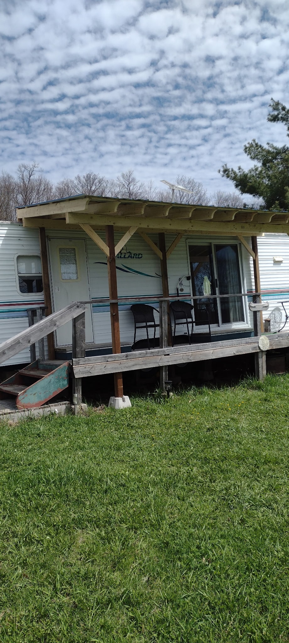 A trailer home is positioned on a grassy area, featuring a front porch supported by wooden beams. Two chairs are placed on the porch, providing a space for relaxation. The exterior displays light-colored siding, with a glass door leading inside.