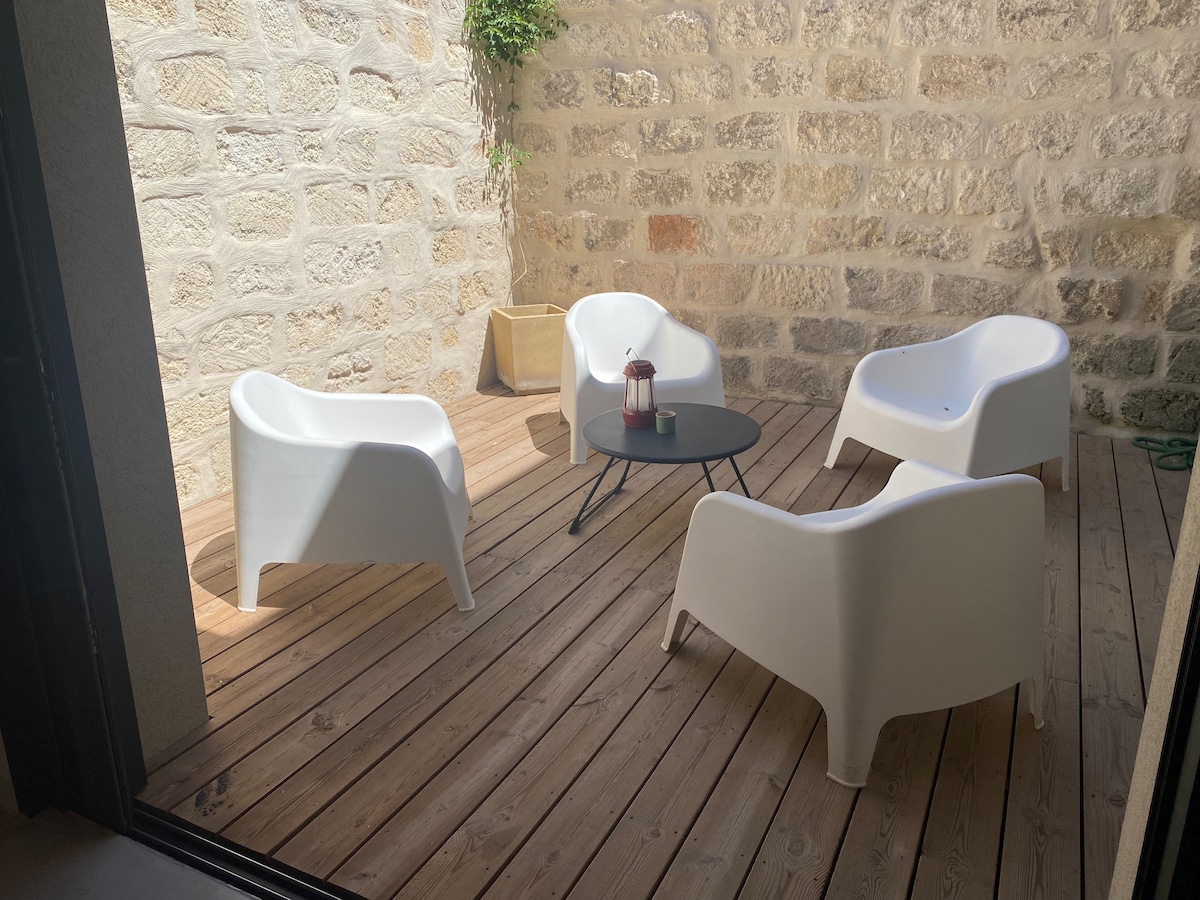 A small outdoor seating area features four white armchairs arranged around a black coffee table. The area is surrounded by a stone wall, offering a secluded atmosphere. Natural wood decking is underfoot, and a small lantern is placed on the table.