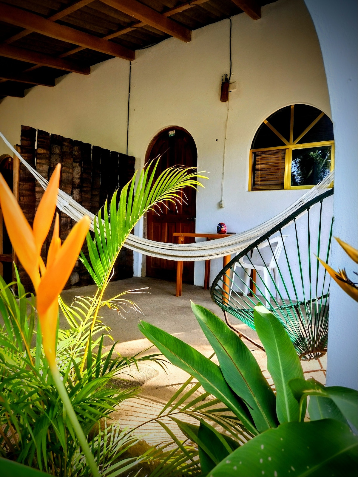 A serene patio area features a gently swaying hammock between two wooden posts. Lush green plants and vibrant orange heliconias frame the space, while a small table is positioned nearby. The arched entrance and windows contribute to a welcoming ambiance.