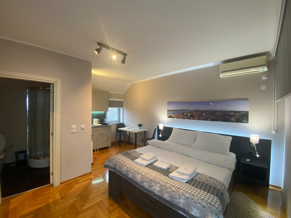 A spacious studio room features a king size bed with crisp white linens and fluffy towels neatly arranged on a gray blanket. A kitchenette is visible in the background, equipped with essential amenities. A glass shower cabin is situated to one side, enhancing the functionality of the space.