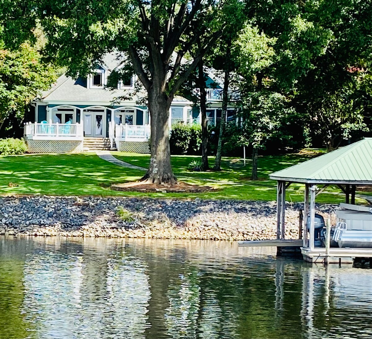 A charming lakefront home is set against a backdrop of lush greenery. The two-story structure features a welcoming porch and large windows that reflect the surrounding trees. A private dock extends into the calm water, providing easy access for boating and relaxation.