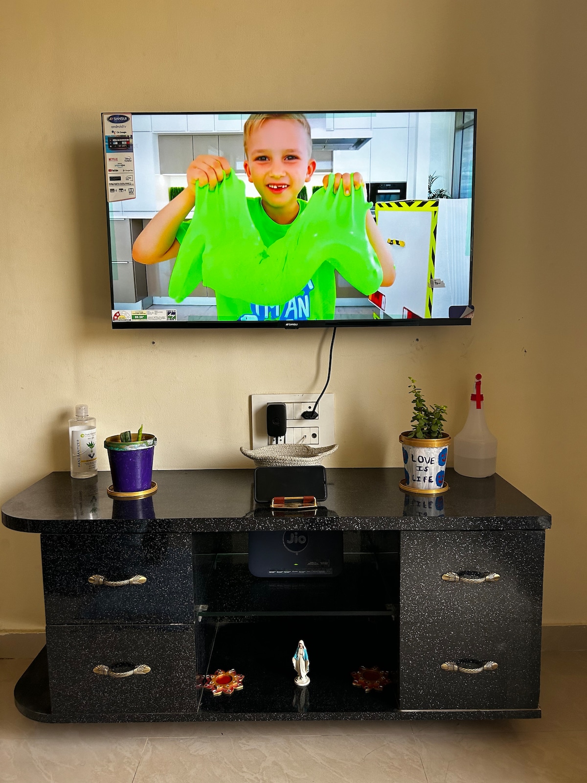 A sleek black TV stand is positioned beneath a mounted flat-screen television, which displays a colorful scene. The stand features decorative elements, including a small potted plant and storage compartments. The light-colored wall provides a neutral backdrop.